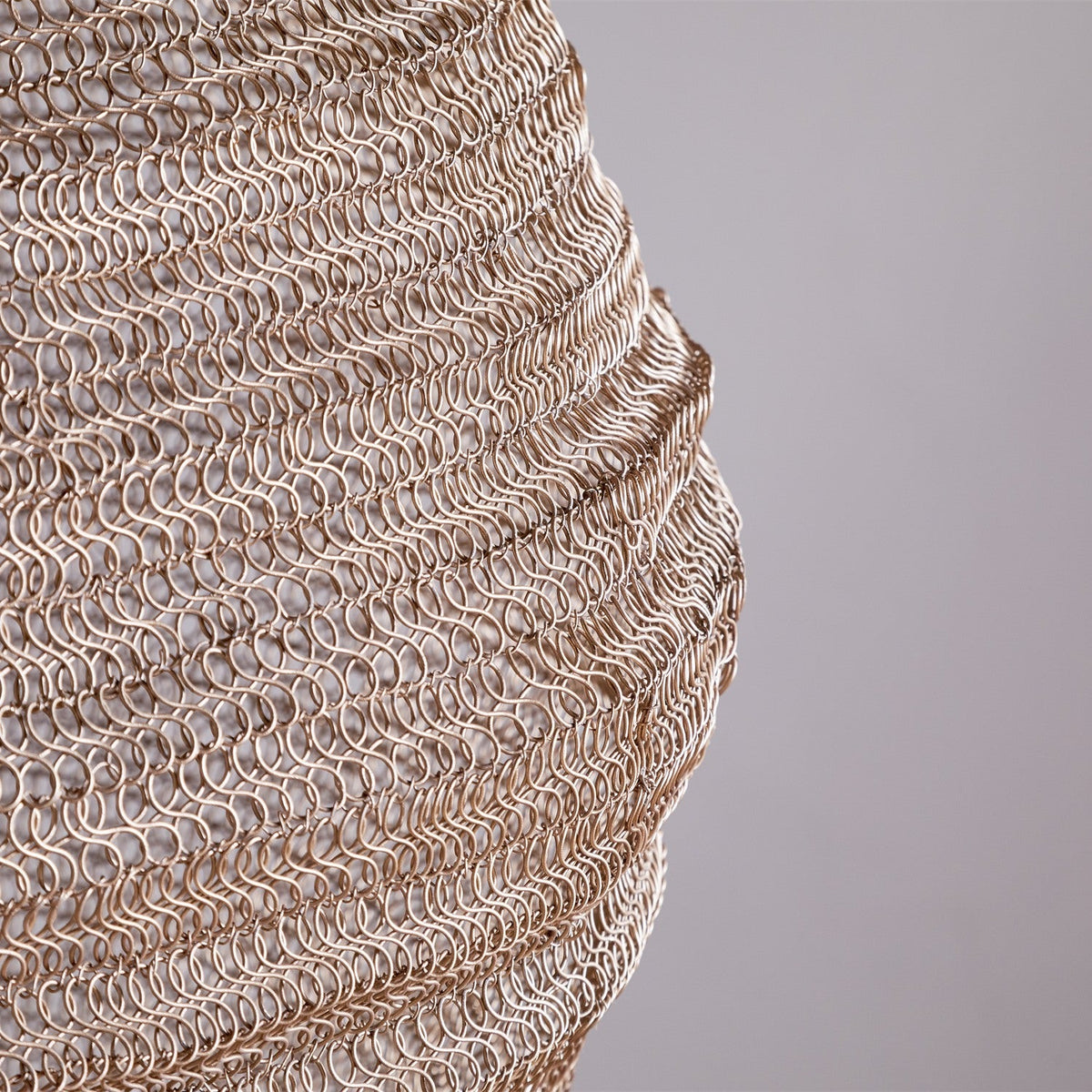 Close-up of the Mukuni Large Wire Lampshade in brass, showing its knitted mesh of interlocking wire loops forming horizontal ridged bands, against a plain light grey background.