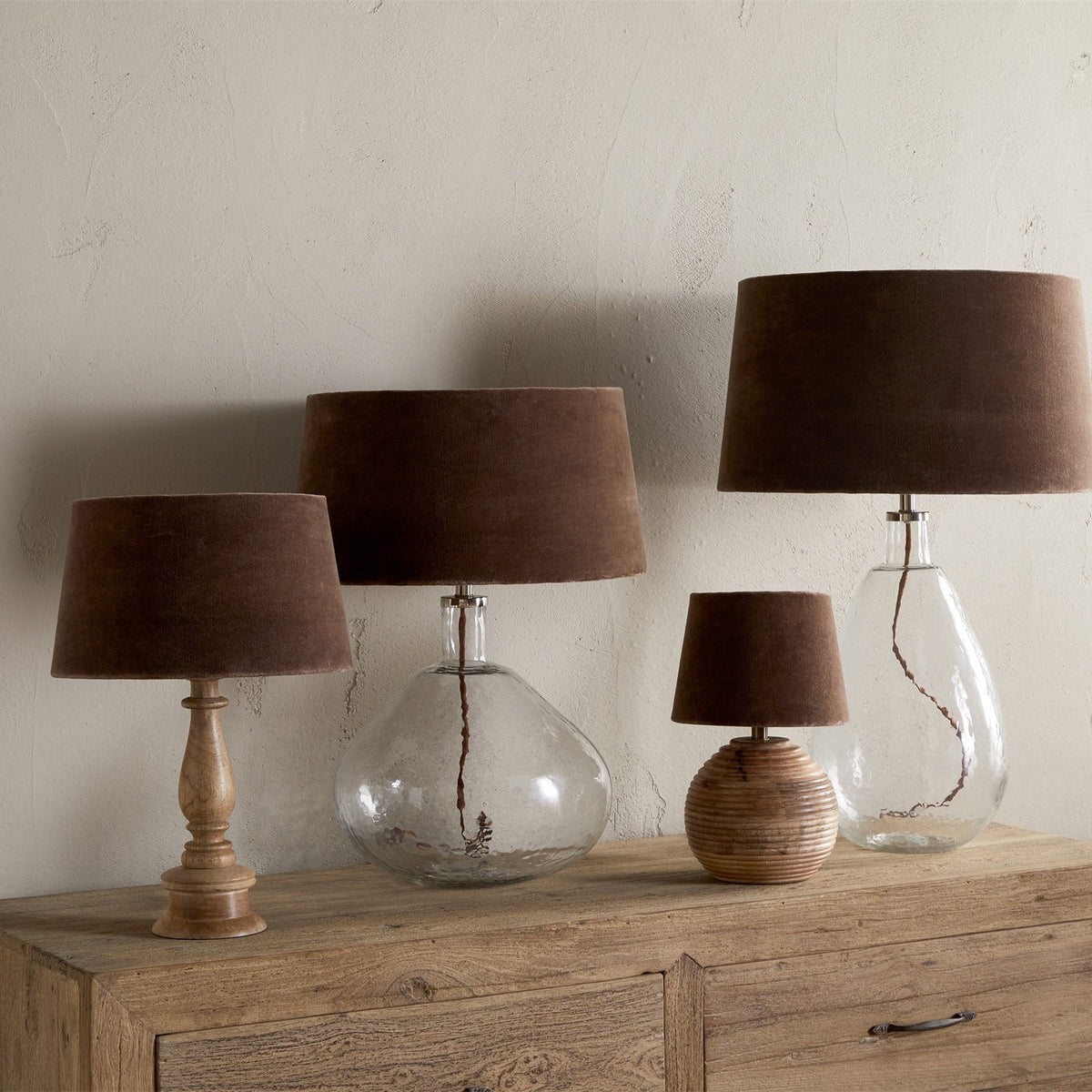 Four table lamps with mocha velvet tapered drum lampshades on wooden, glass, and turned bases, shown on a sideboard against a plain wall.