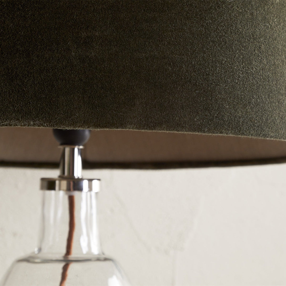 Close-up of the Mendai Medium lampshade in moss green velvet, showing the fitter with chrome gallery and finial atop a clear glass table lamp base.