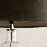 Close-up of the Mendai Medium lampshade in moss green velvet, showing the fitter with chrome gallery and finial atop a clear glass table lamp base.
