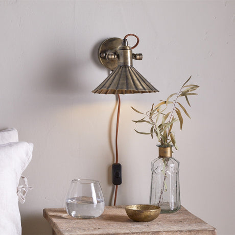 Moti Wall Light in antique brass with a pleated scallop-edge shade on a jointed arm, plug-in cord with switch, shown above a bedside table in a bedroom setting.