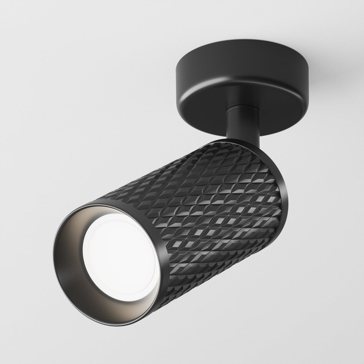 The Focus Design Single Spotlight - Black is a modern, black cylindrical track light with a textured diamond pattern, mounted on a white ceiling. It features a GU10 base for efficient illumination and emits a bright glow.