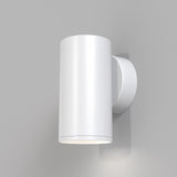 The FOCUS S Down Wall Light - White is a sleek cylindrical sconce on a light gray wall. With a minimalist design, its smooth surface emits soft downward light, casting gentle shadows that enhance any modern industrial look.