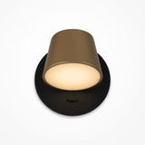 A modern lamp with a cylindrical gold shade and a sleek black & brass circular base. When on, it emits a soft, warm glow with adjustable lighting options. The background is plain white.