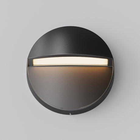 The Mane Outdoor LED Round Wall Light features a black finish and modern design, casting efficient downward light. Its curved top offers a semi-circular glow below, seamlessly blending with the light gray wall for an elegant outdoor illumination.