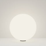 The 49cm Erda Pedestal Light in white, with its modern design and illuminated round form, emits a soft glow on a plain surface, enhancing outdoor lighting with understated charm.