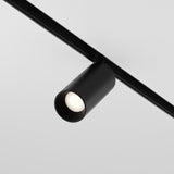 The Focus LED Magnetic Exility Track Spotlight 5W 3000K - Black, with its cylindrical design, mounts on a ceiling track to cast low glare light downward over a light gray backdrop, showcasing its sleek and modern minimalism.