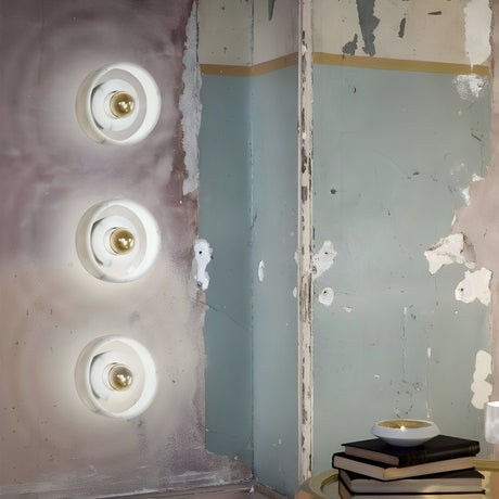 Three Marble Wall Light - White & Gold fixtures are mounted vertically on a distressed, two-toned wall with peeling paint. In the corner, there’s a small table topped with stacked books and a white bowl.