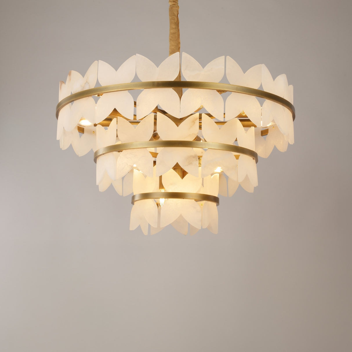 Niori Petraia three-tier chandelier with scalloped alabaster petals on brushed brass rings, suspended from a fabric-wrapped chain against a pale background.