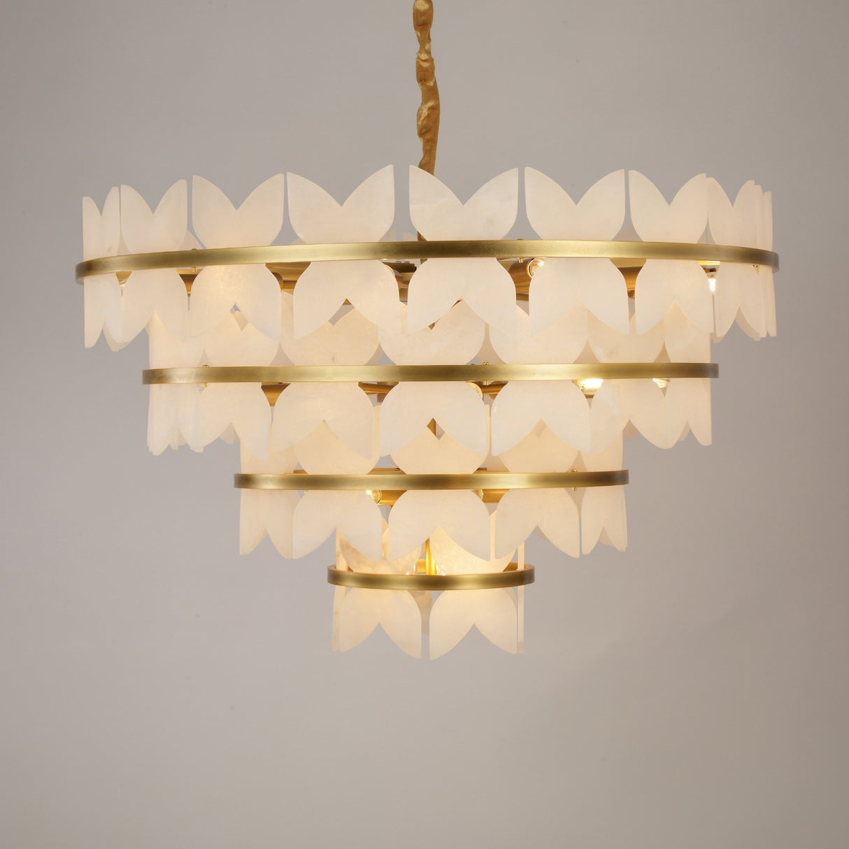 Niori Petraia four-tier chandelier featuring sculpted alabaster petals around brushed brass rings, suspended from a rope-wrapped drop against a grey background.