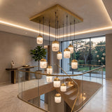 Renzara LED 12 Light Cluster Alabaster Chandelier in brass, twelve capsule shades at varied drop heights from a square canopy, shown above a staircase landing.