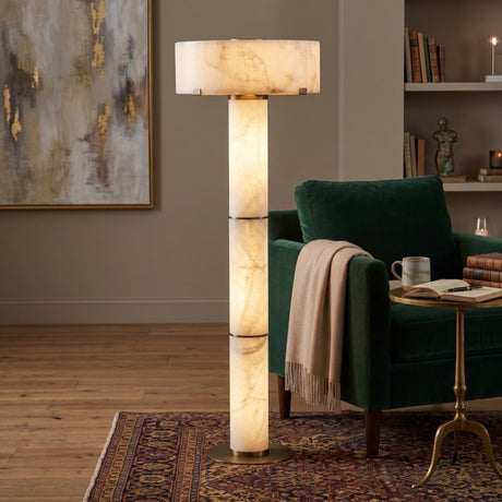 Colonnetta 3 Light Column Alabaster Floor Lamp with brushed brass collars and base disc, glowing beside a green armchair in a living room setting.
