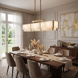 Quadrena Linear Alabaster Chandelier 113cm in Brushed Brass, glowing rectangular bar with two-tier stone-effect panels, shown above a dining table in a neutral-toned room.
