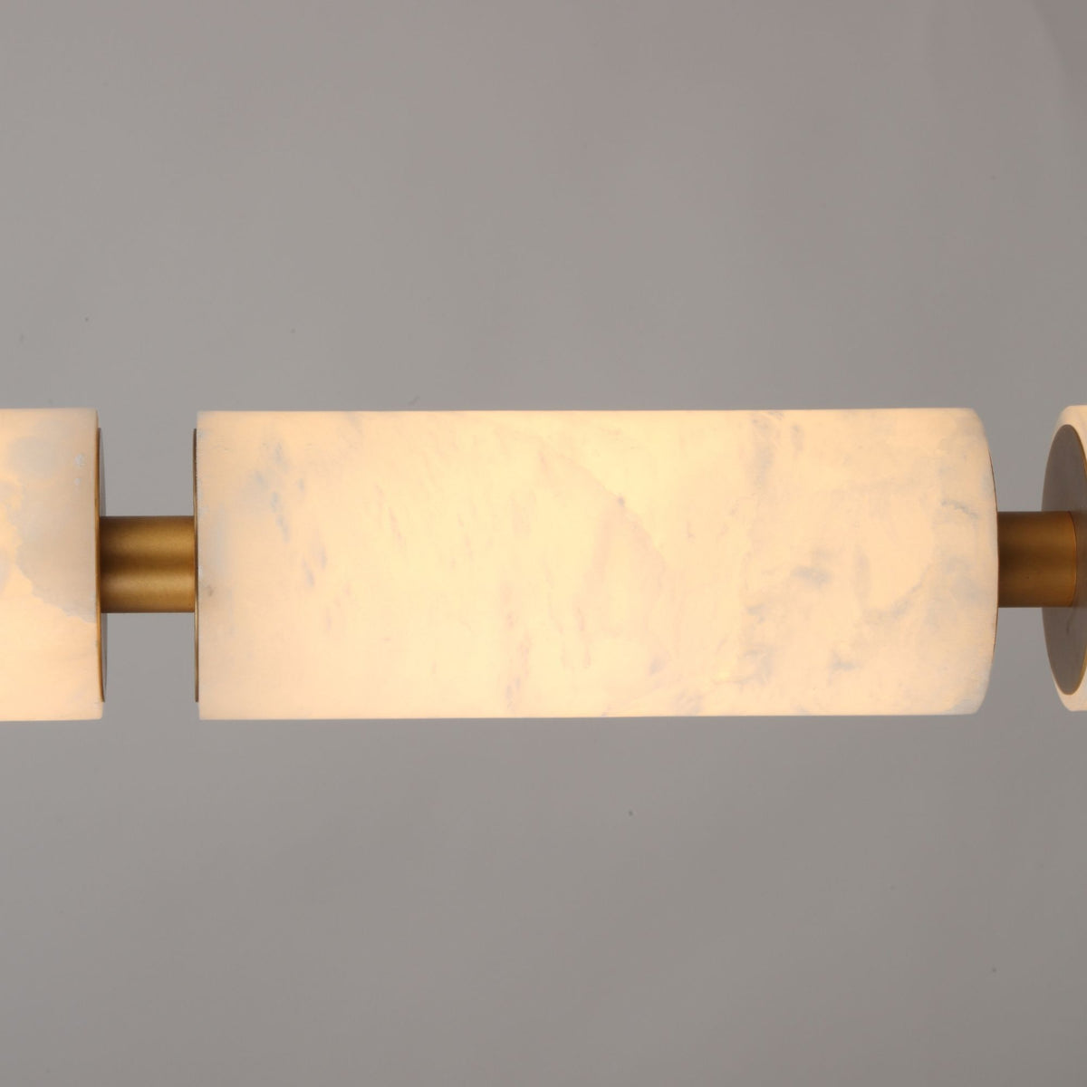 Close-up of the Ardent LED alabaster column pendant light, showing natural stone cylinders joined by brushed brass spacers, glowing warm white against a plain grey background.