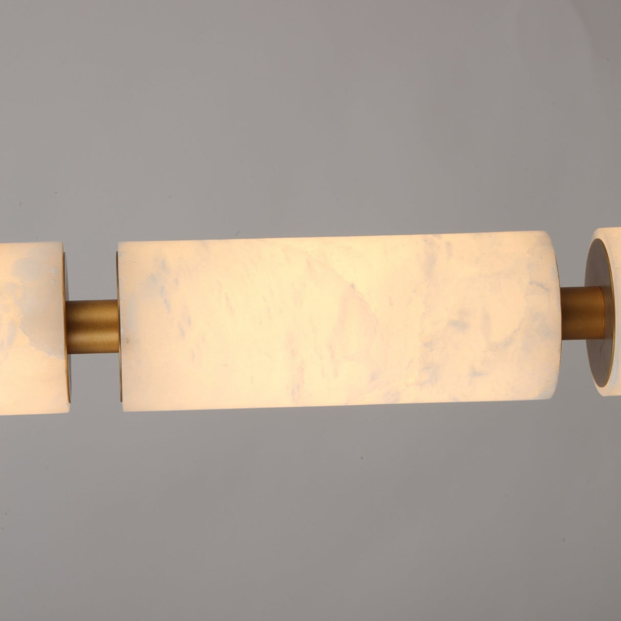 Close-up of Niori Ardent Medium alabaster column pendant light, showing veined white stone cylinders joined by a brushed brass collar, illuminated softly.