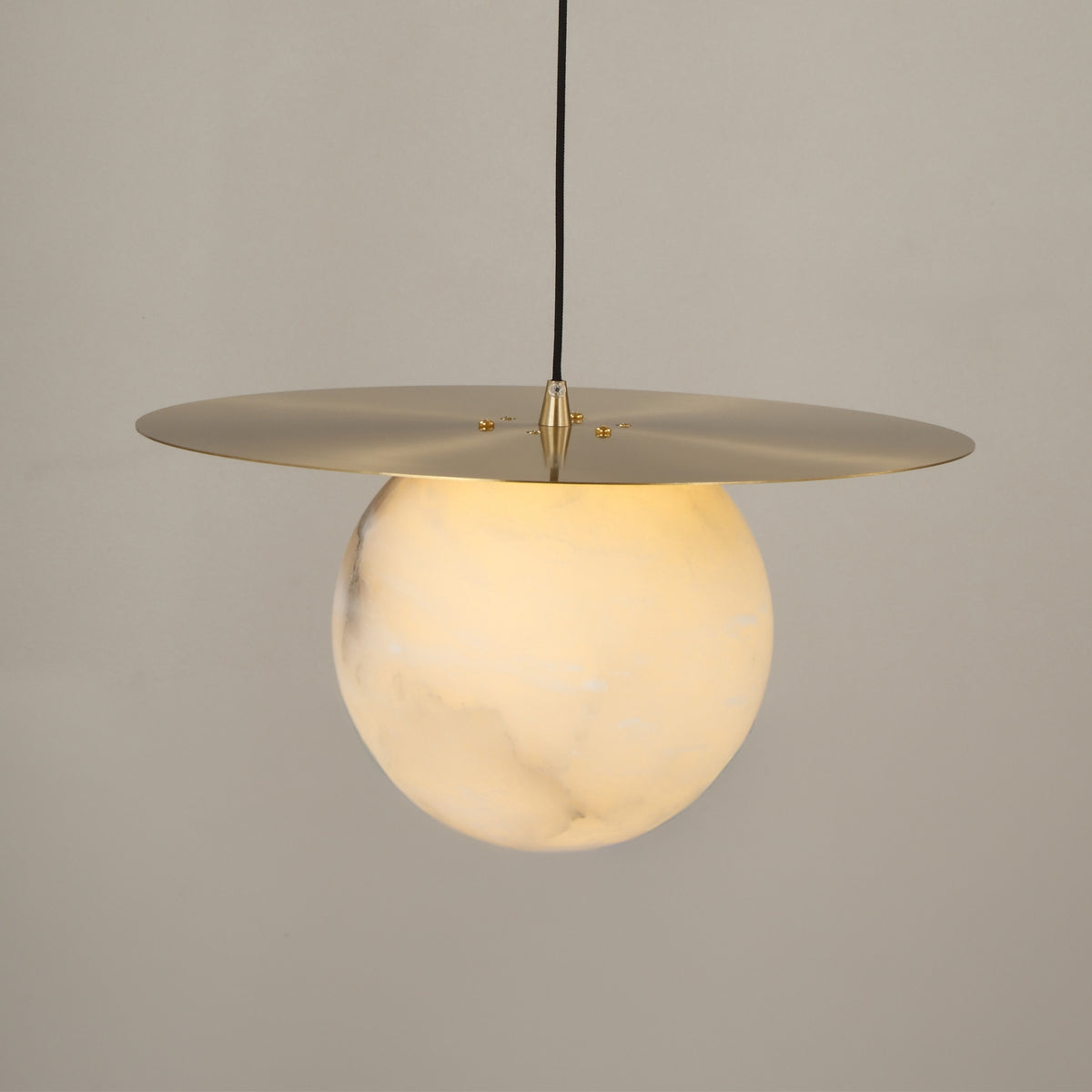 Marenzo Small Orb Alabaster Pendant Light in Brushed Brass, showing a spherical white alabaster shade beneath a flat brass ceiling disc, lit against a plain grey background.