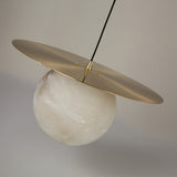 Marenzo Alabaster Orb Pendant Light in Brushed Brass, featuring a natural alabaster sphere suspended beneath a flat brass reflector disc, shown against a plain light grey background.