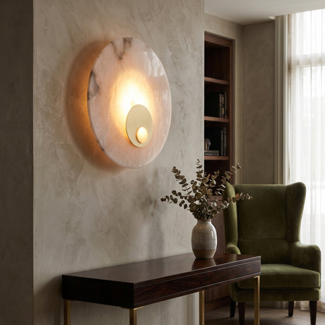 Selvana LED Small Alabaster Wall Light in soft white, round natural stone disc with brushed brass centre boss, glowing warmly on a plaster wall in a living room setting.