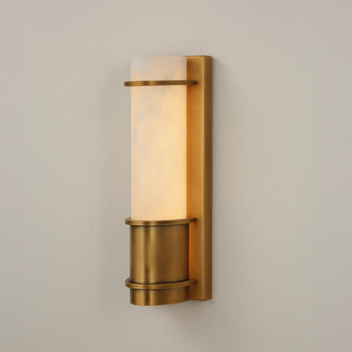 Niori Averna Small LED wall light with a cylindrical Spanish alabaster column set on a brushed brass backplate with banded cuff, against a light grey wall.