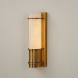 Niori Averna Small LED wall light with a cylindrical Spanish alabaster column set on a brushed brass backplate with banded cuff, against a light grey wall.