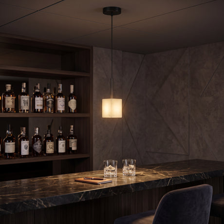 Draxen Alabaster Drum Pendant Light in matte black and brass, glowing warmly above a marble bar counter with whisky glasses and shelved bottles.