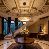 Bacinella LED bowl alabaster pendant light in brushed brass, hung above a round wooden table in an entrance hall with a domed ceiling and curved staircase.