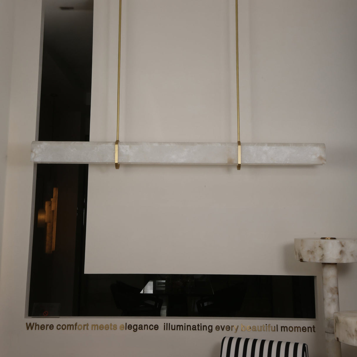 Niori Zarune LED linear chandelier with a horizontal Spanish alabaster bar suspended by two brushed brass drop rods in a contemporary showroom setting.