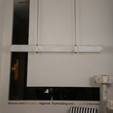 Niori Zarune LED linear chandelier with a horizontal Spanish alabaster bar suspended by two brushed brass drop rods in a contemporary showroom setting.