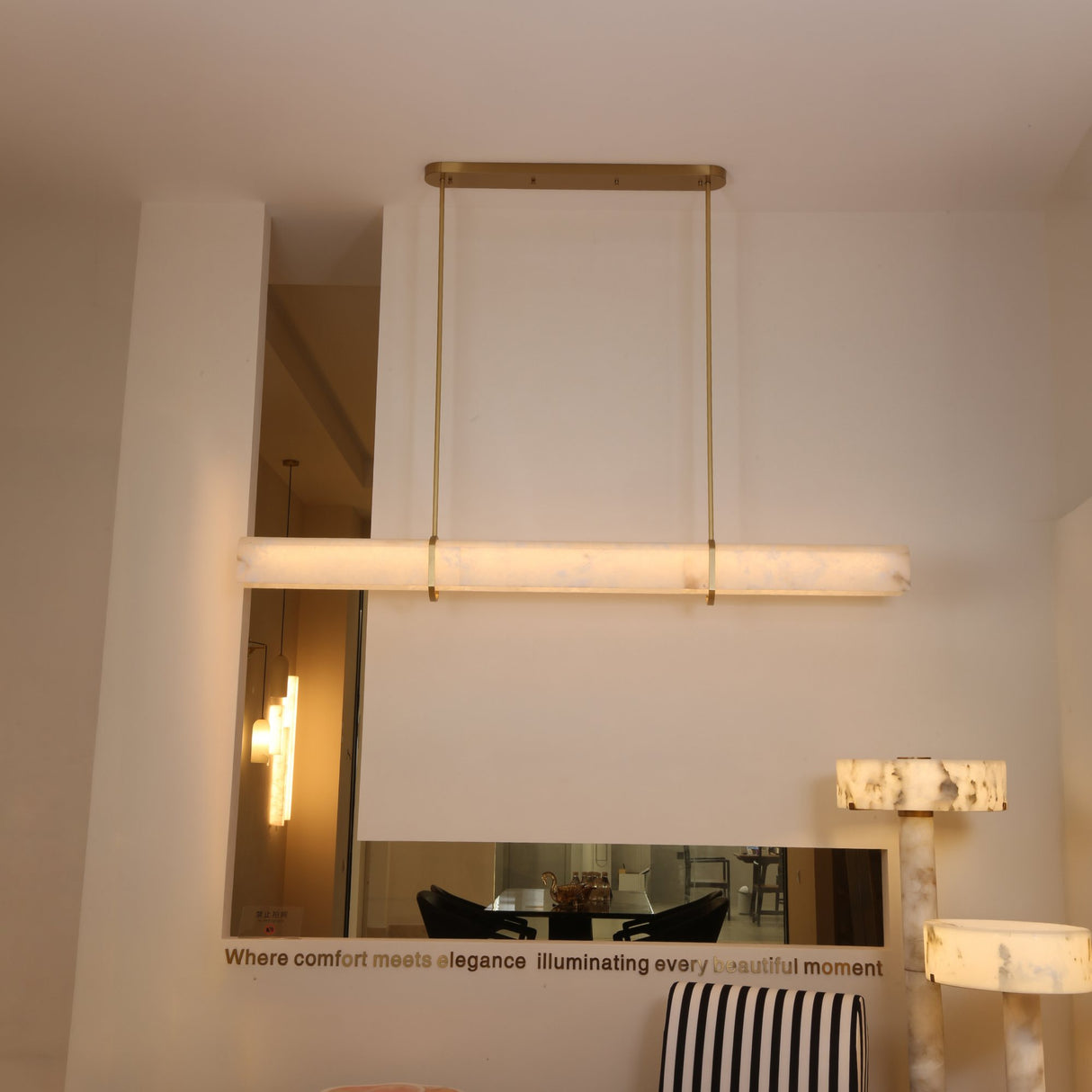 Niori Zarune LED linear alabaster chandelier in brushed brass, suspended from an oval canopy by two slim rods in a contemporary showroom setting.