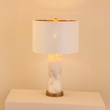 Calix Alabaster Pillar Table Lamp with white fabric drum shade, brushed brass base and finial, lit against a plain warm background.