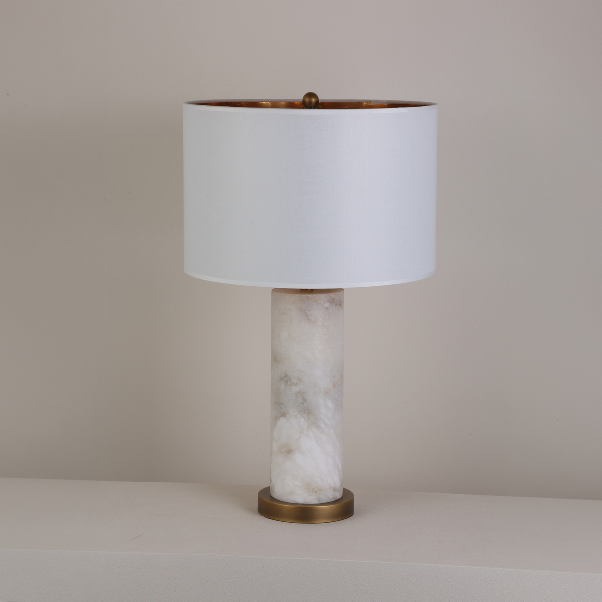Calix Alabaster Pillar Table Lamp with brushed brass base and finial, white fabric drum shade with gold-toned interior, on a plain light background.