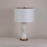 Calix Alabaster Pillar Table Lamp with brushed brass base and finial, white fabric drum shade with gold-toned interior, on a plain light background.