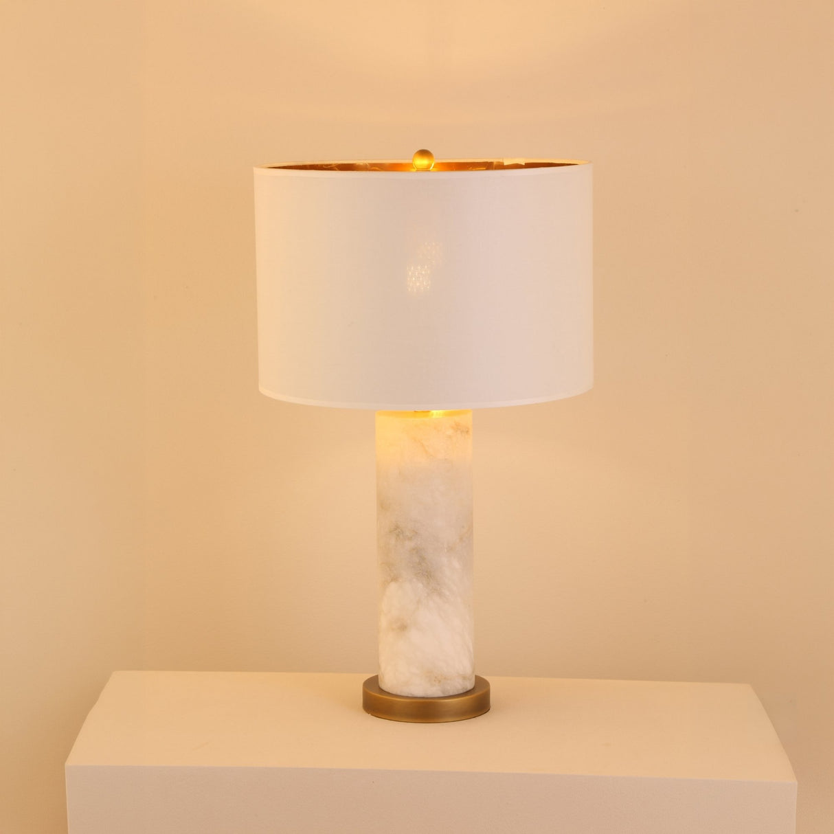 Calynor 1 Light Column Alabaster Table Lamp with white marbled stone base, brushed brass stem, and ivory drum shade, glowing softly against a plain light background.