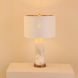 Calynor 1 Light Column Alabaster Table Lamp with white marbled stone base, brushed brass stem, and ivory drum shade, glowing softly against a plain light background.