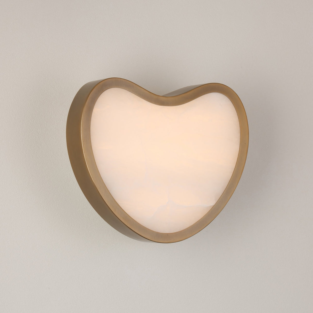 Niori Marea LED wall light with a heart-shaped brushed brass shell framing a natural Spanish alabaster face, glowing warm white against a light grey wall.