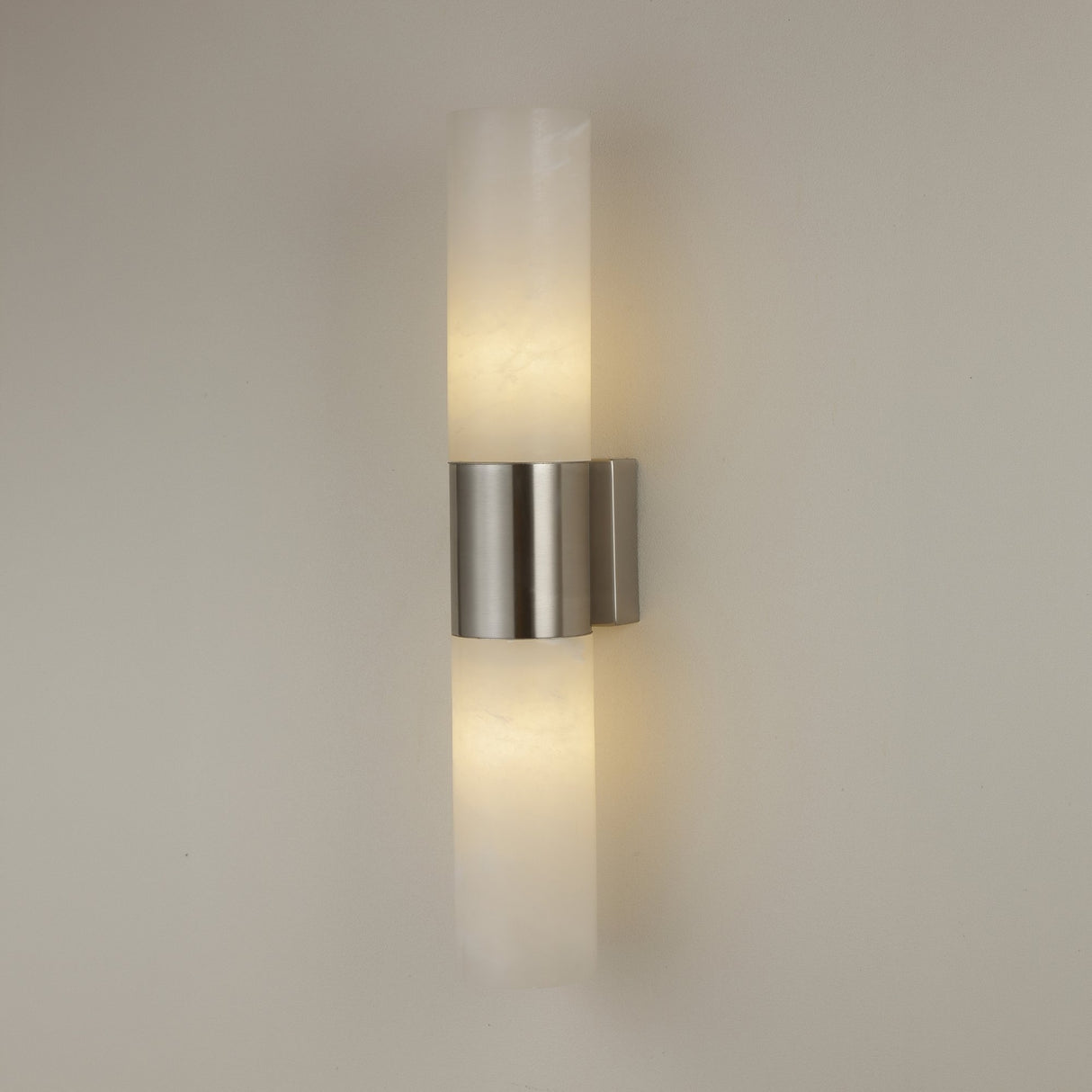 Niori Fiora LED alabaster wall light with two vertical stone cylinders joined by a satin nickel band, casting warm light on a neutral wall.