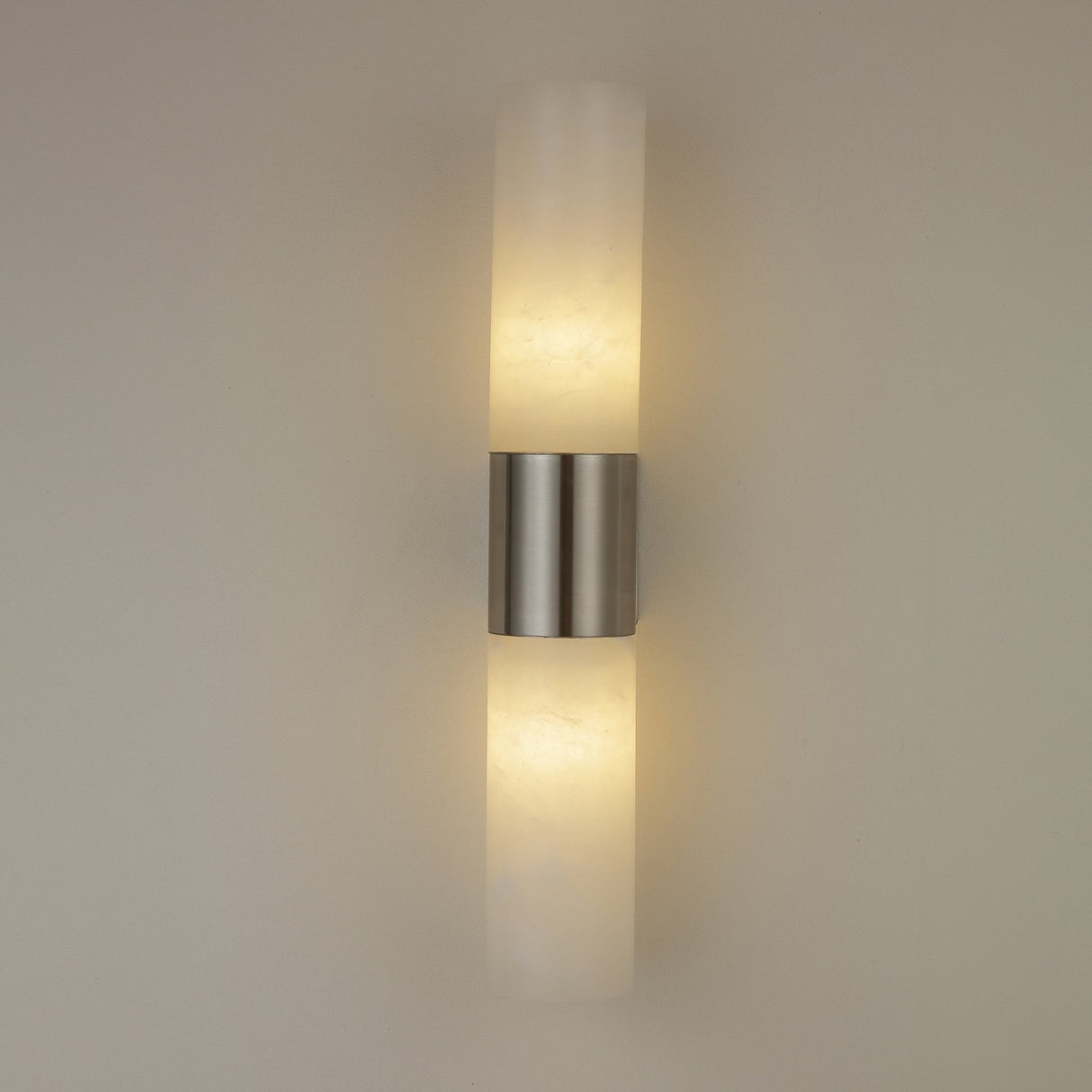 Niori Fiora LED wall light illuminated on a neutral wall, showing two alabaster cylinders glowing warm white above and below a satin nickel band.