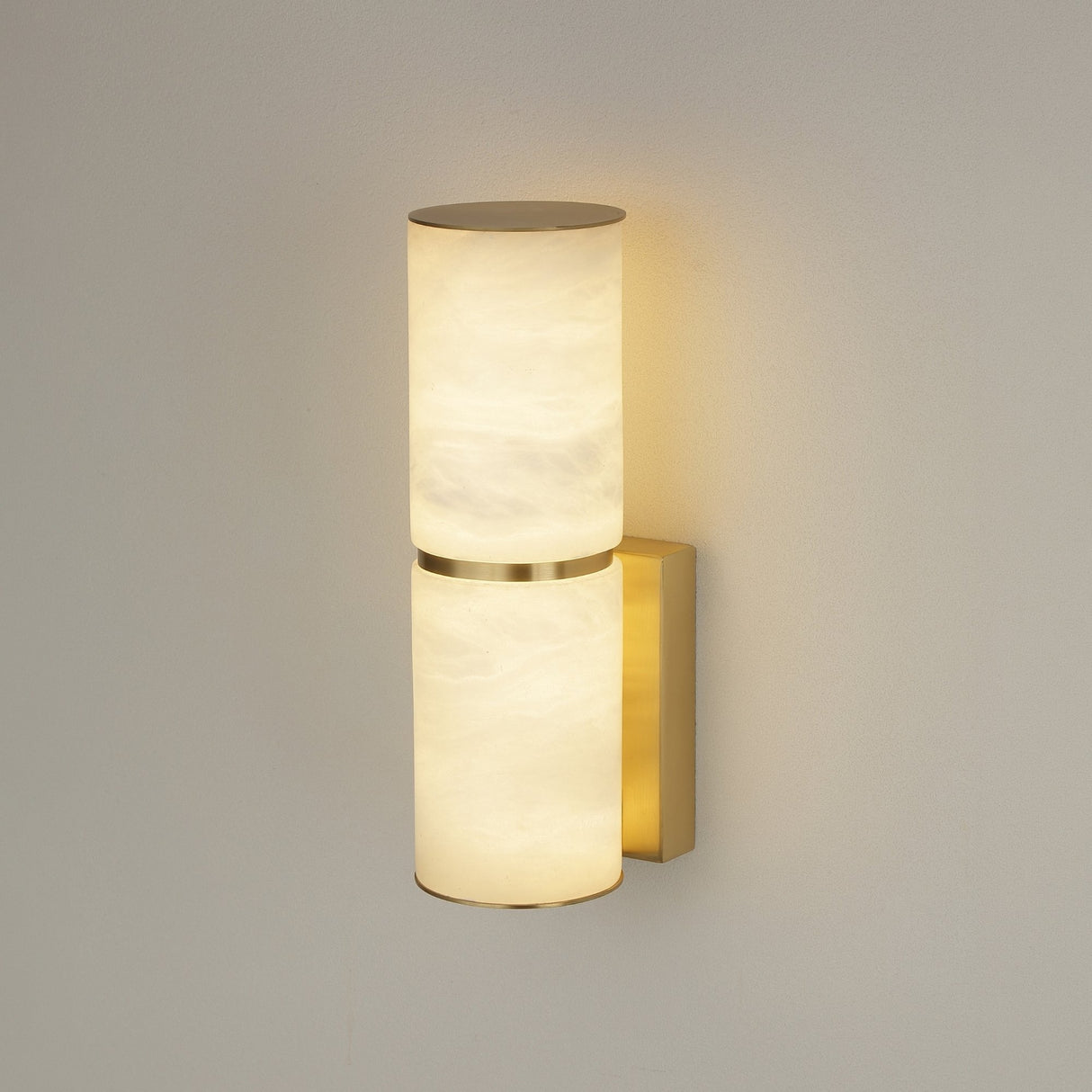Niori Almora Small wall light with two stacked Spanish alabaster cylinders, brushed brass band and backplate, illuminated warm white against a pale wall.