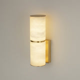 Niori Almora Small wall light with two stacked Spanish alabaster cylinders, brushed brass band and backplate, illuminated warm white against a pale wall.