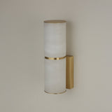 Niori Almora Small LED wall light with two stacked Spanish alabaster cylinders, brushed brass band, trims, and rectangular backplate on a pale wall.