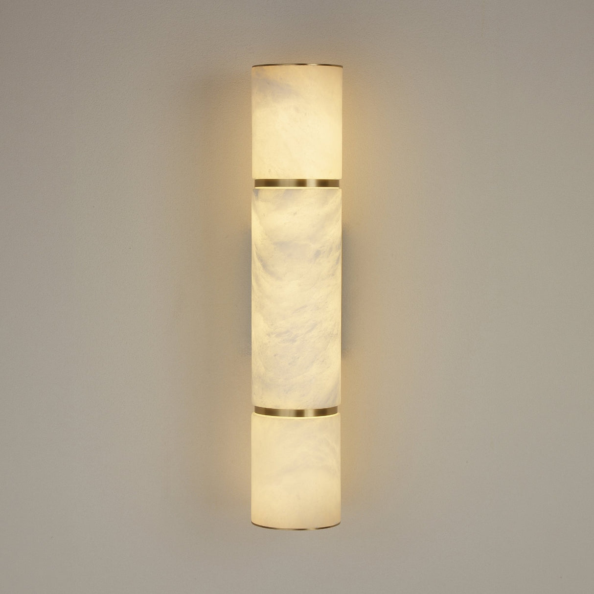 Niori Almora LED alabaster cylinder wall light in brass, illuminated with warm up and down glow, mounted on a light grey wall.
