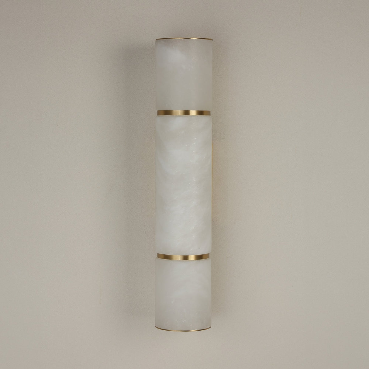 Niori Almora medium wall light with a natural Spanish alabaster cylinder segmented by brass bands, mounted against a plain neutral wall.