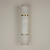 Niori Almora medium wall light with a natural Spanish alabaster cylinder segmented by brass bands, mounted against a plain neutral wall.