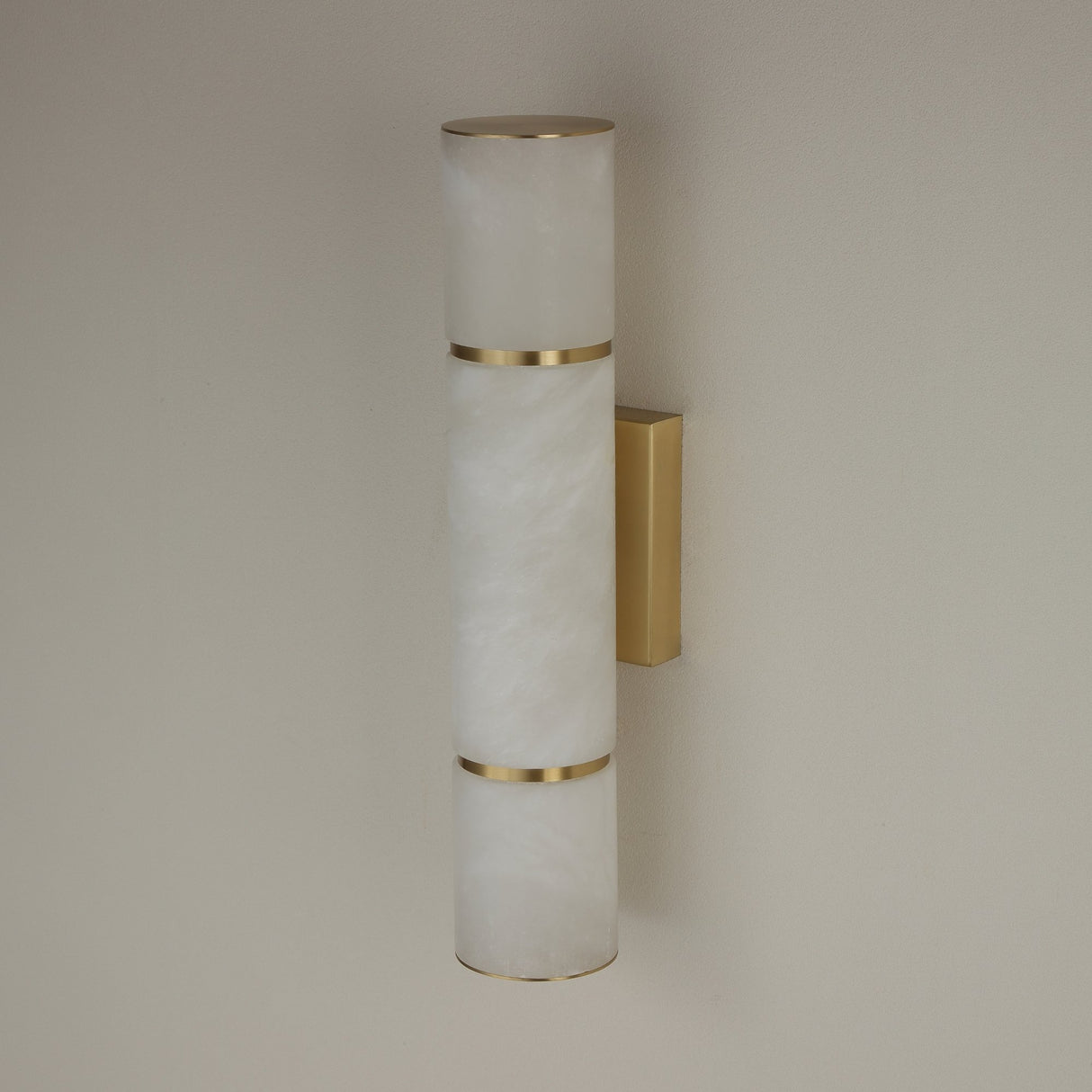 Niori Almora LED alabaster cylinder wall light in a medium size, with brass banding and rectangular brushed brass wall plate on a neutral wall.