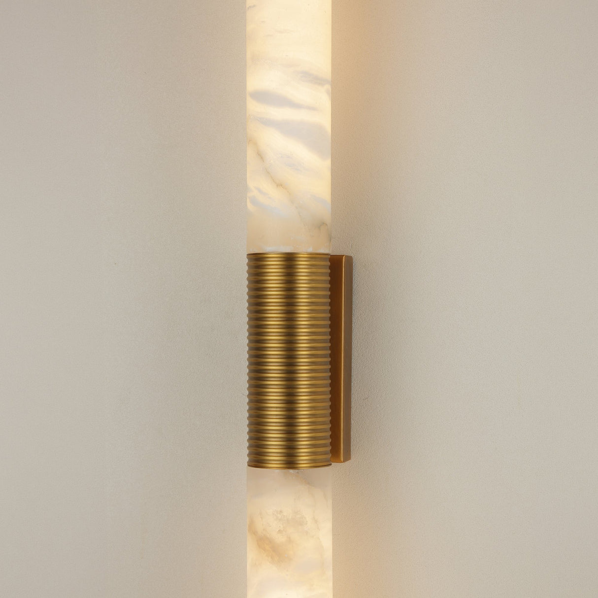 Eryon LED Large alabaster wall light with two cylindrical stone columns joined by a brushed brass mount and ribbed bronze centre sleeve, illuminated.