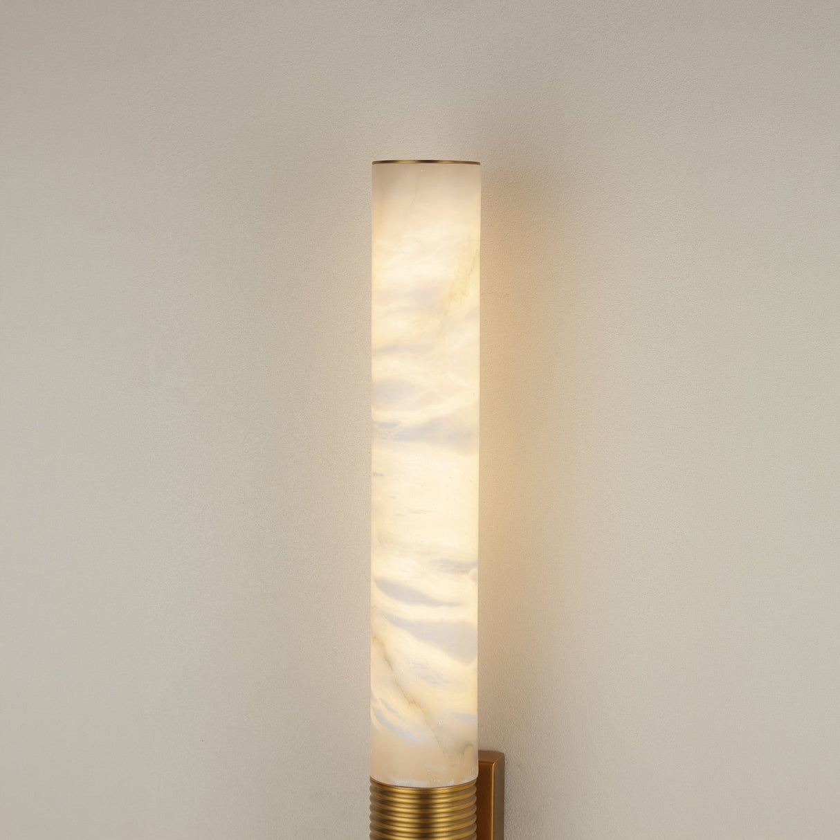 Eryon LED large alabaster wall light illuminated on a pale wall, showing the tall cylindrical stone column above a brushed brass ribbed mount.