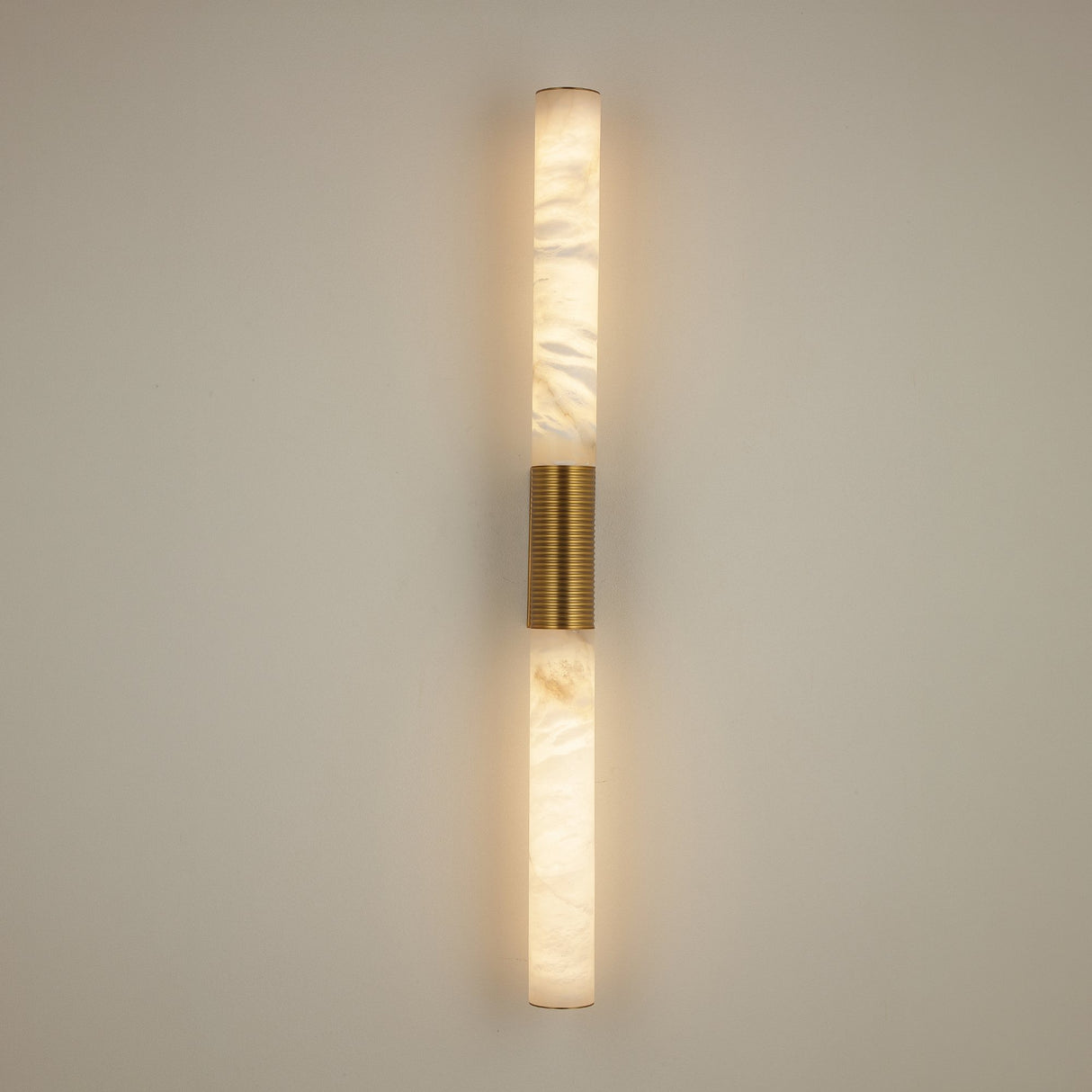 Niori Eryon LED large wall light with two natural alabaster cylinders joined by a brushed brass and ribbed bronze centre mount, illuminated on a pale wall.
