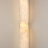 Vera Small Silhouette alabaster wall light, slim vertical column with an hourglass waist and brushed brass detail, glowing warm white against a plain grey wall.