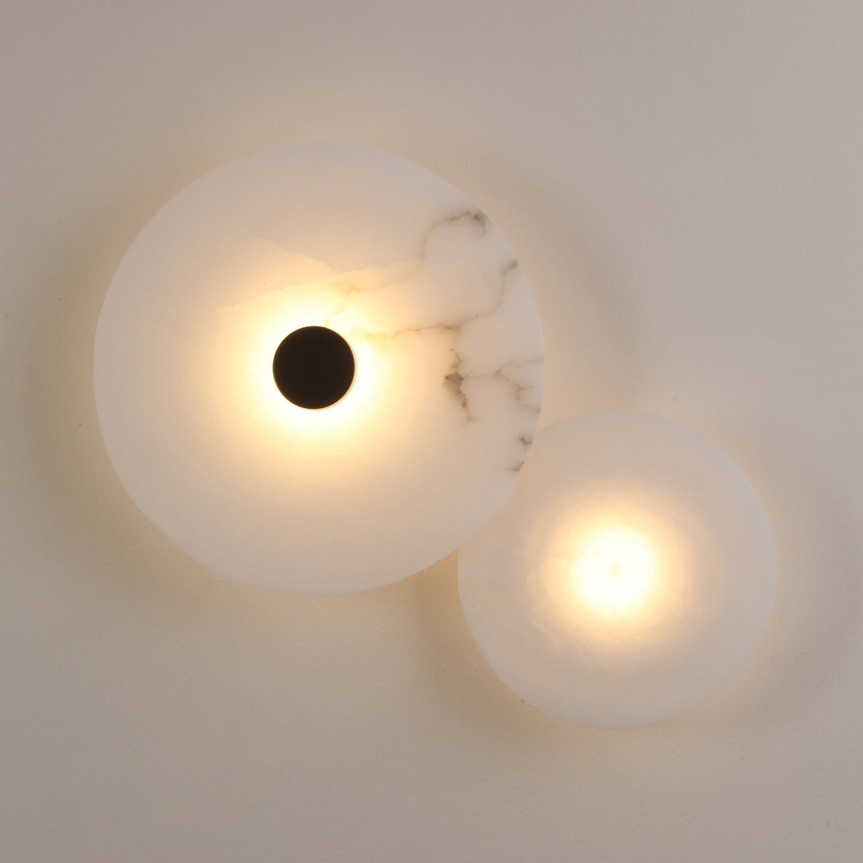 Solmira LED Dual Disc Alabaster Wall Light with overlapping large and small alabaster discs, brushed brass centre detail, and warm white glow, mounted on a plain light wall.
