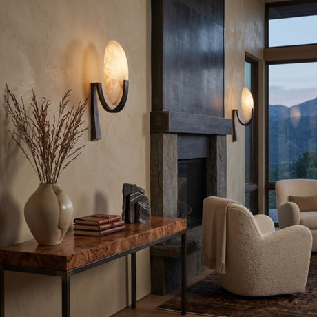 Mica 1 Light Disc Alabaster Wall Light in matt black, illuminated on a living room wall beside a stone fireplace with mountain views.